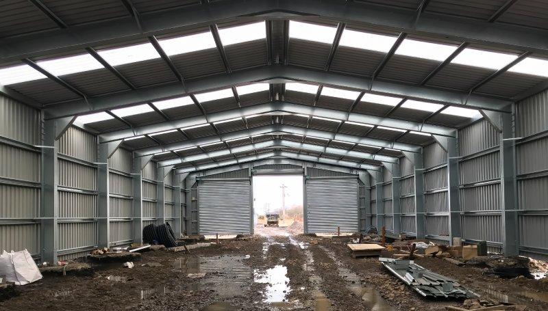Internal view of a large-scale steel warehouse construction with skylight panels and a muddy floor.
