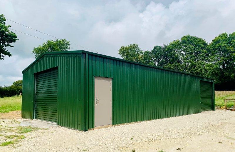 A long forest green corrugated metal warehouse with a front-facing roller door and a side personal access entrance.