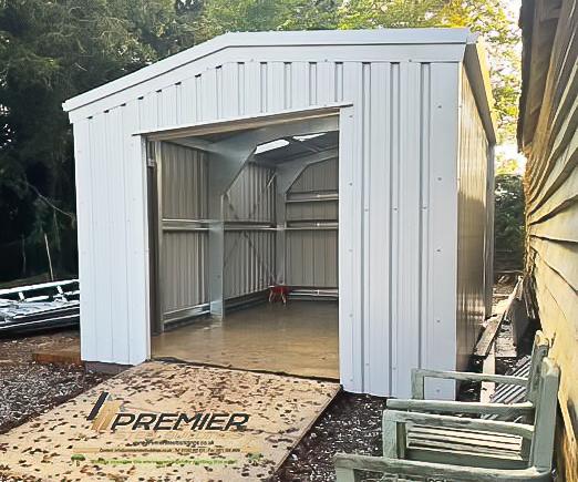 Front view of a light-colored steel shed with the roller door open, revealing the internal structure and concrete floor.